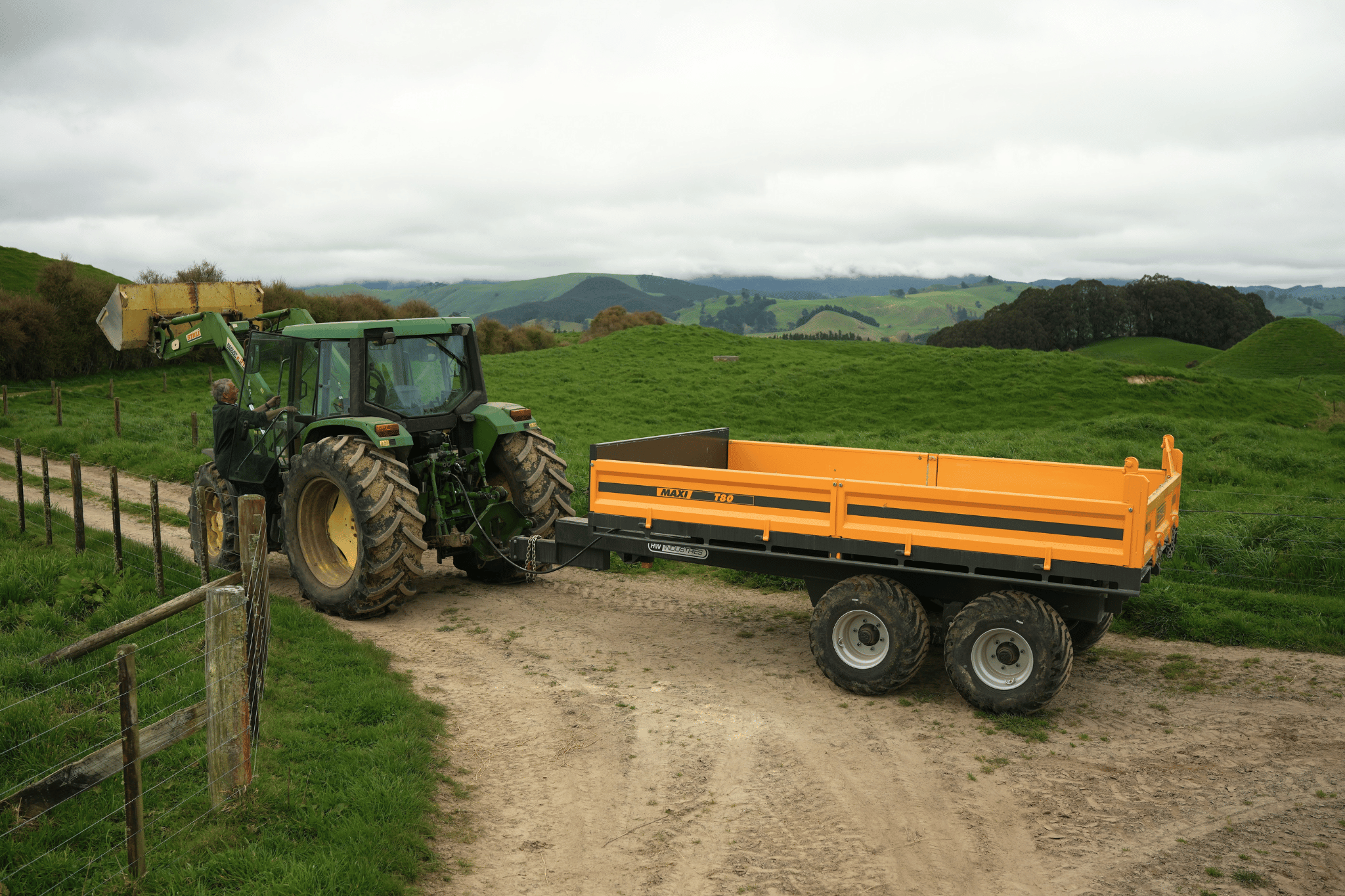 Farmer climbing into a John Deere tractor towing an orange Maxi 180 tip trailer on a hill-country track