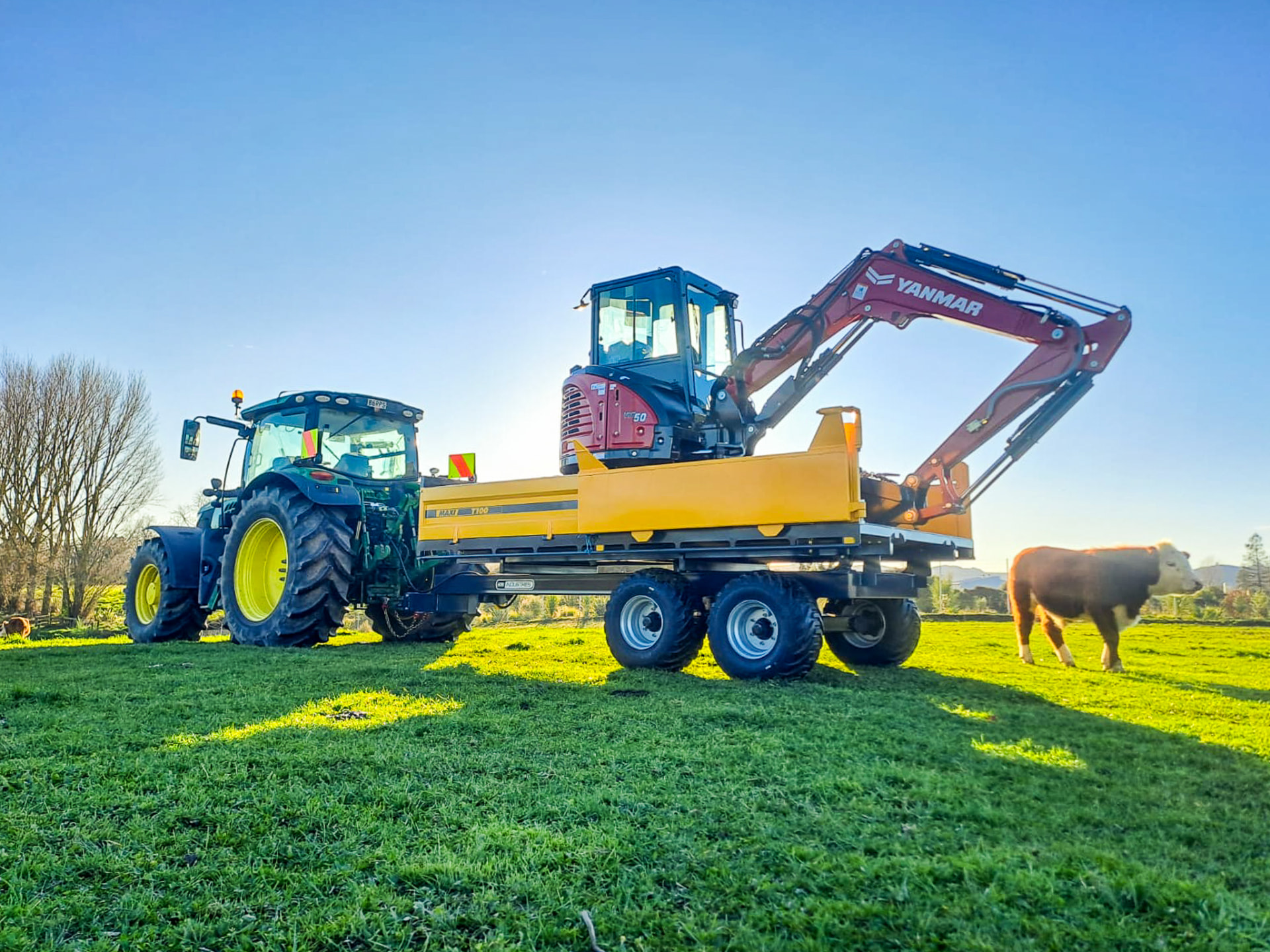 Maxi T100 tip trailer carrying a Yanmar excavator behind a tractor on a farm paddock